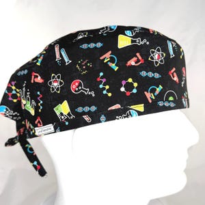May include: Black surgical scrub cap with colorful science-themed illustrations. The cap features images of atoms, DNA strands, microscopes, and beakers. The cap has a tie closure and a small tag that reads "LoveNstitches".