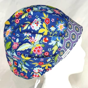 May include: A blue scrub cap featuring a vibrant floral pattern with red, yellow, and light blue flowers and green leaves. The front band displays a geometric hexagonal print in purple, blue, and white. A small white tag reads LoveNstitches.