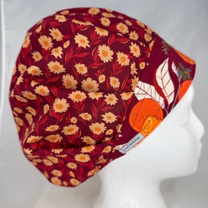 May include: A scrub cap featuring a deep burgundy background with a repeating pattern of light orange daisies and red leaves. The front band displays a vibrant print of sliced oranges and green foliage, with a small white tag reading LoveNatitchies.