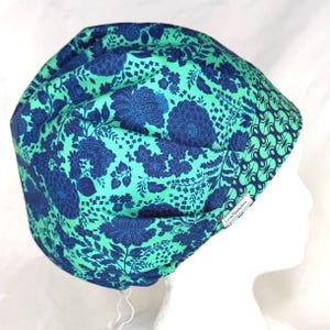 May include: A scrub cap featuring a vibrant mint green background with a dark blue floral pattern. The cap includes a contrasting band with a dark blue and mint green geometric print and a small white tag labeled LoveNotitches.