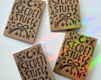 May include: Four small, rectangular notebooks with the words "SECRET STUFF" in black, stylized lettering. The covers are a light brown color and feature a crescent moon, stars, a key, and floral designs. The notebooks are arranged on a white surface.
