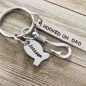 May include: A silver keychain with a fish hook charm and a fish-shaped tag engraved with "Jackson." The rectangular tag reads "Hooked on Dad." The keychain is on a wooden surface.