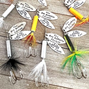 May include: A collection of seven silver fishing lures with colorful feathers and a triple hook. Each lure has a silver tag with the text "The Bob Baptiste Classic" and "Lake Shasta" followed by a year and a name.