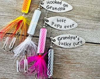 Könnte beinhalten: Fünf Angelköder mit unterschiedlich gefärbten Federn und Körpern. Jeder Köder hat ein Metalletikett mit einer personalisierten Botschaft: "best Daddy ever", "Hooked on Grandpa", "best Papa ever" und "Grandpa's lucky lure".