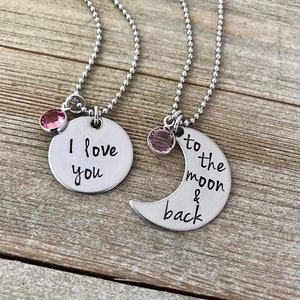 Puede incluir: Dos collares de plata con dijes a juego. Un dije es un c&iacute;rculo con el texto "Te amo" y el otro es una luna creciente con el texto "hasta la luna y de regreso". Ambos dijes tienen un peque&ntilde;o cristal rosa.