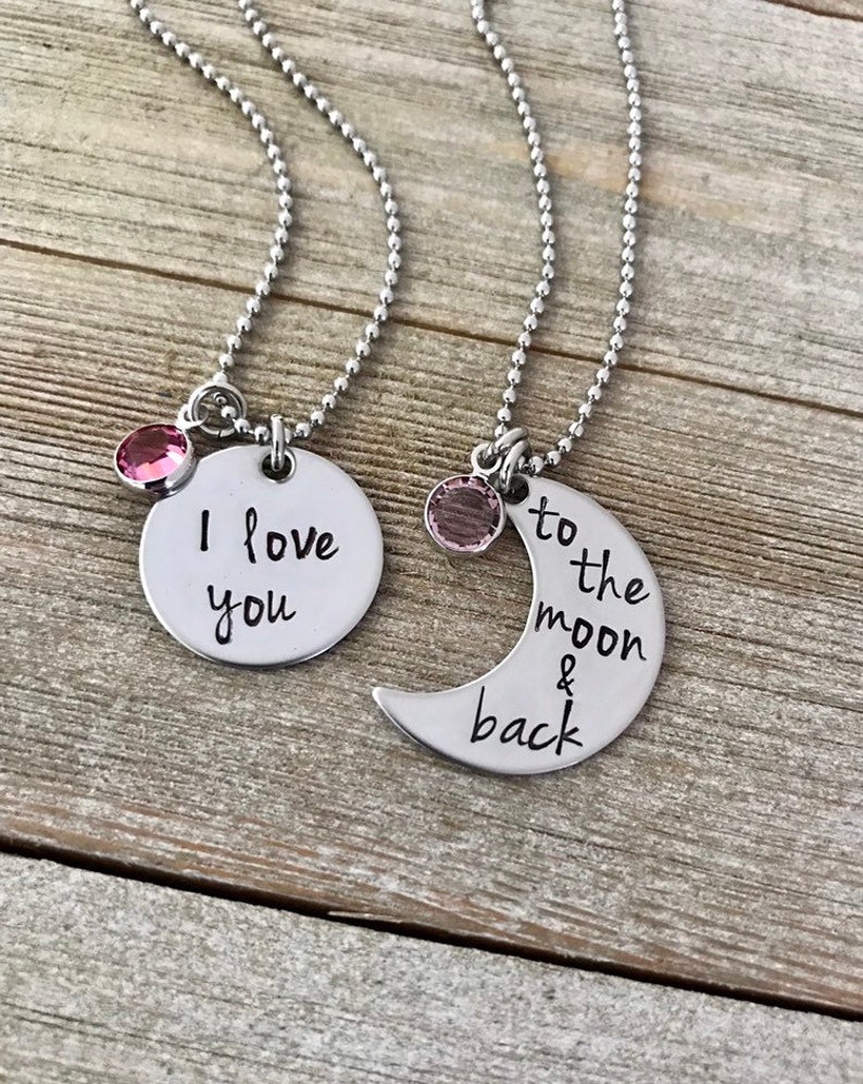 Puede incluir: Dos collares de plata con dijes a juego. Un dije es un c&iacute;rculo con el texto "Te amo" y el otro es una luna creciente con el texto "hasta la luna y de regreso". Ambos dijes tienen un peque&ntilde;o cristal rosa.
