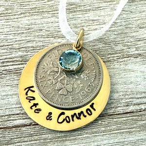 May include: A gold-toned pendant necklace with a silver coin and a blue gemstone. The pendant features the names "Kate & Connor" and a coin dated 1963. The necklace hangs from a white ribbon.