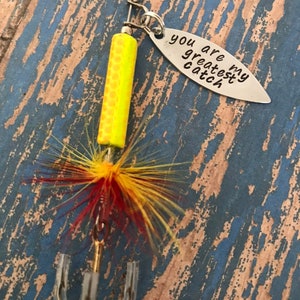 May include: A silver fishing lure with a yellow and orange body, red and yellow feathers, and a silver tag that reads "You are my greatest catch".