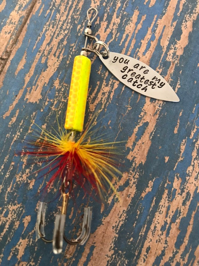May include: A silver fishing lure with a yellow and orange body, red and yellow feathers, and a silver tag that reads "You are my greatest catch".