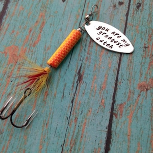 May include: A silver fishing lure with a yellow and orange body and red and yellow feathers. The lure has a silver oval tag with the text "You are my greatest catch".