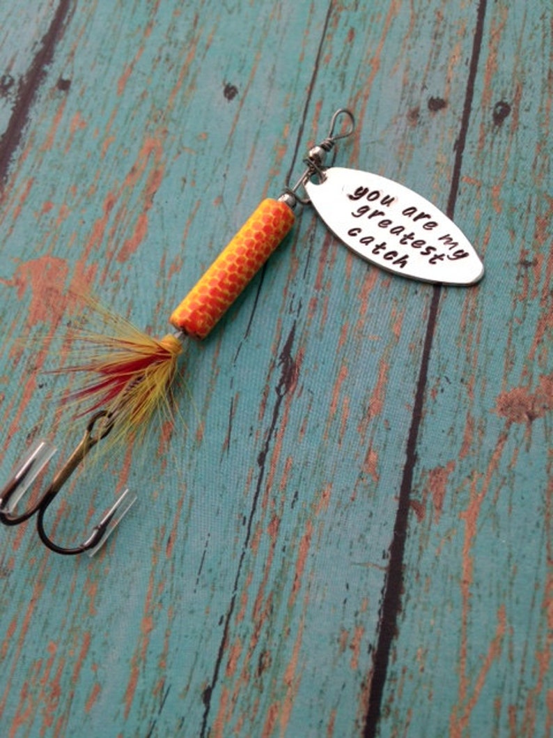 May include: A silver fishing lure with a yellow and orange body and red and yellow feathers. The lure has a silver oval tag with the text "You are my greatest catch".