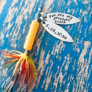 May include: A silver fishing lure with a yellow and orange body and red and yellow feathers. The lure has two tags attached to it. One tag says "You are my greatest catch" and the other tag says "5.28.2016".