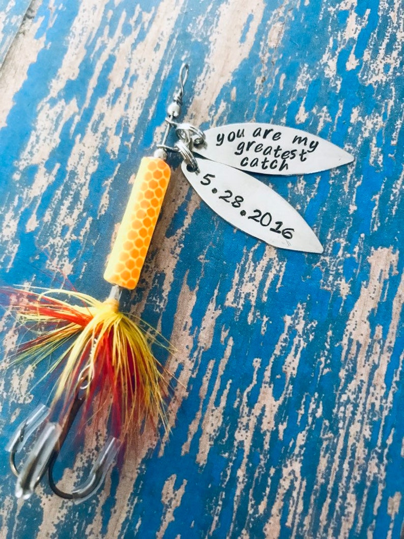 May include: A silver fishing lure with a yellow and orange body and red and yellow feathers. The lure has two tags attached to it. One tag says "You are my greatest catch" and the other tag says "5.28.2016".