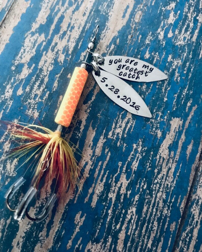 May include: A silver fishing lure with a yellow and orange body and a red and yellow feather tail. The lure has two tags attached to it. The top tag says "You are my greatest catch" and the bottom tag says "5.28.2016".