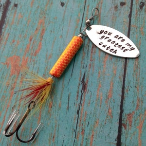 May include: A silver fishing lure with a yellow and orange body and red and yellow feathers. The lure has a silver oval tag that says "You are my greatest catch."