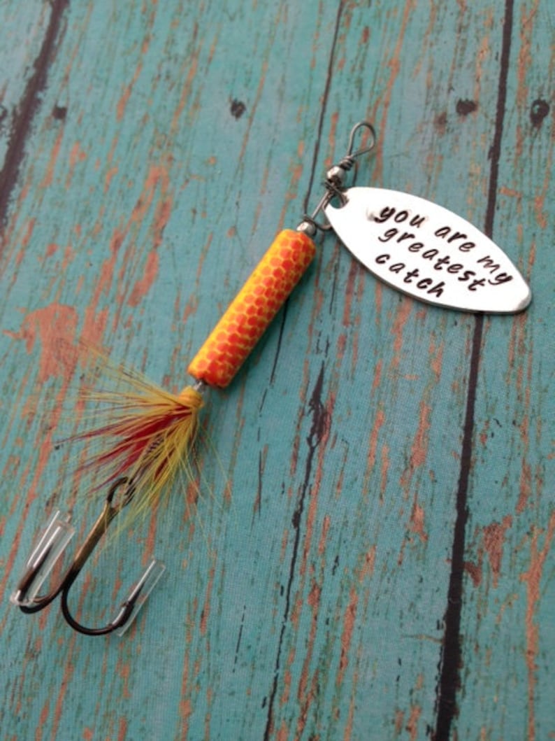 May include: A silver fishing lure with a yellow and orange body and red and yellow feathers. The lure has a silver oval tag that says "You are my greatest catch."