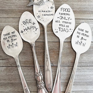 May include: A collection of five vintage silver spoons with stamped text and designs. One spoon features a mermaid, another says "Bee Happy," and others read "Frog Parking Only" and "Thank You for Helping Me Grow."