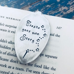 May include: A silver spoon bookmark with the text "Create your own Story" and a star design. The spoon is lying on a book with the text "147 aid the" visible.
