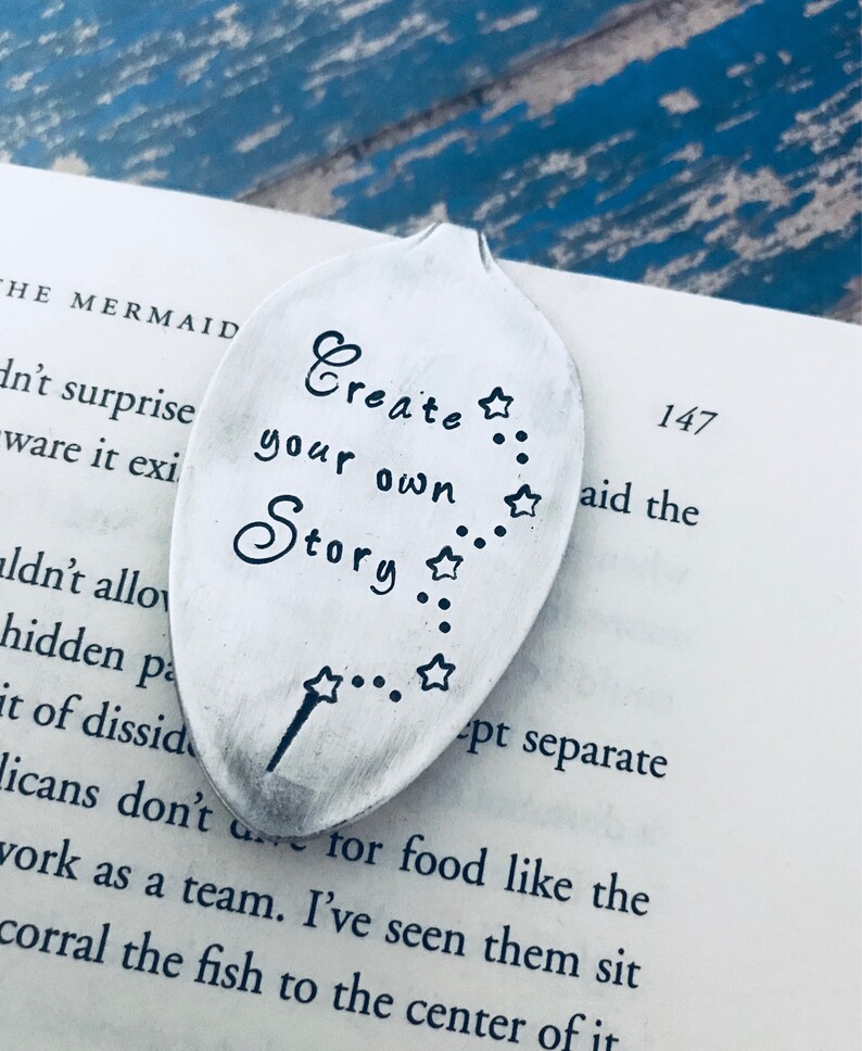 May include: A silver spoon bookmark with the text "Create your own Story" and a star design. The spoon is lying on a book with the text "147 aid the" visible.