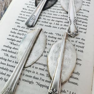 May include: Three silver spoons with ornate handles, repurposed as bookmarks. The spoons are placed on a page of a book with text visible.