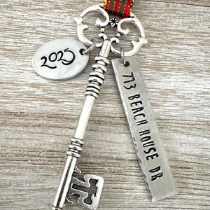 May include: A silver key-shaped ornament with a decorative top and a tag that reads "713 BEACH HOUSE DR." An oval tag displays the year "2025." The ornament hangs from a red, green, and gold striped ribbon.
