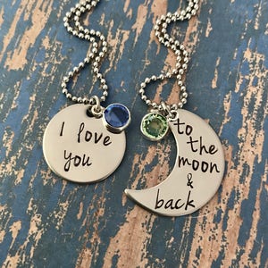Puede incluir: Dos collares de plata con colgantes a juego. Un colgante es un c&iacute;rculo con las palabras "Te amo" grabadas en &eacute;l y una piedra preciosa azul. El otro colgante es una luna creciente con las palabras "hasta la luna y de regreso" grabadas en &eacute;l y una piedra preciosa verde.