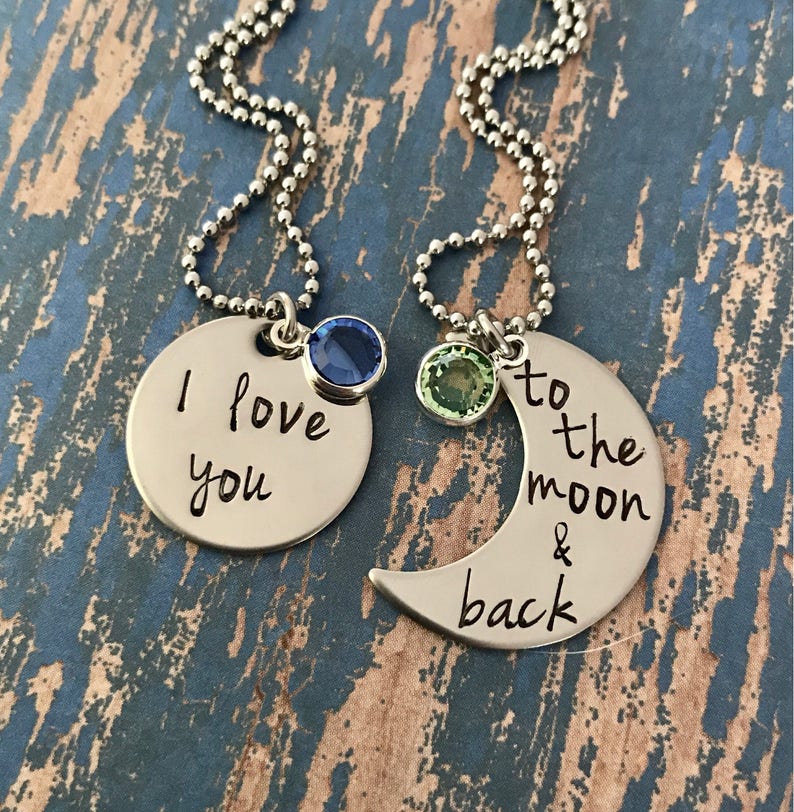 Puede incluir: Dos collares de plata con colgantes a juego. Un colgante es un c&iacute;rculo con las palabras "Te amo" grabadas en &eacute;l y una piedra preciosa azul. El otro colgante es una luna creciente con las palabras "hasta la luna y de regreso" grabadas en &eacute;l y una piedra preciosa verde.