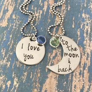 Puede incluir: Dos collares de plata con dijes. Un dije dice "Te amo" con una piedra preciosa azul. El otro dije es una luna creciente con el texto "hasta la luna y de regreso" y una piedra preciosa verde.