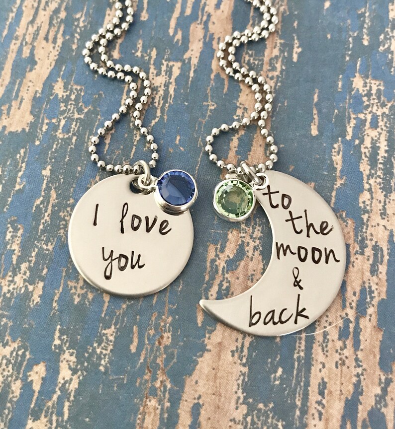 Puede incluir: Dos collares de plata con dijes. Un dije dice "Te amo" con una piedra preciosa azul. El otro dije es una luna creciente con el texto "hasta la luna y de regreso" y una piedra preciosa verde.
