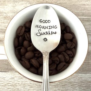 May include: A white coffee mug with a gold rim filled with dark brown coffee beans. A silver spoon rests inside the mug, engraved with the words "Good Morning Sunshine" and a small flower.