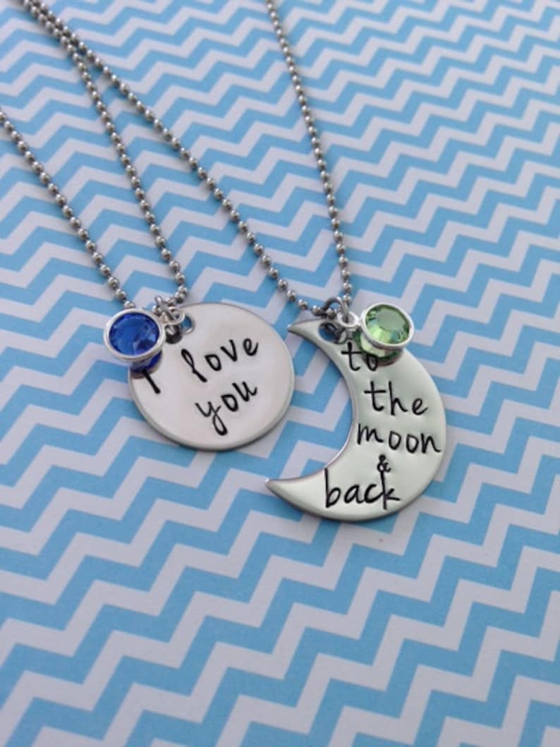 Puede incluir: Dos collares de plata con dijes. Un dije es un c&iacute;rculo con el texto "Te amo" y un cristal azul. El otro dije es una luna creciente con el texto "hasta la luna y de regreso" y un cristal verde.
