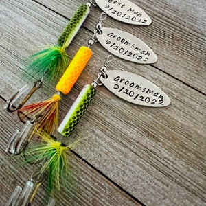Pu&ograve; includere: Tre esche da pesca con etichette personalizzate. Le etichette sono argentate e riportano il testo "Best Man 9/10/2022", "Groomsman 9/10/2022" e "Groomsman 9/10/2022". Le esche sono verdi, gialle e arancioni.