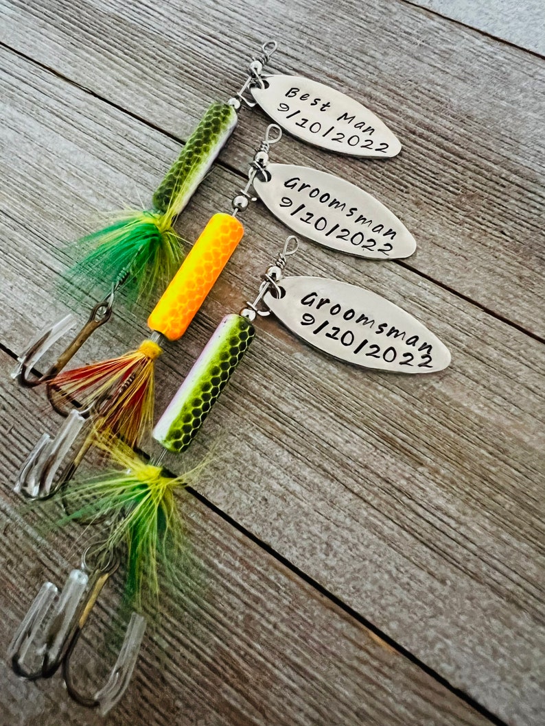 Pu&ograve; includere: Tre esche da pesca con etichette personalizzate. Le etichette sono argentate e riportano il testo "Best Man 9/10/2022", "Groomsman 9/10/2022" e "Groomsman 9/10/2022". Le esche sono verdi, gialle e arancioni.