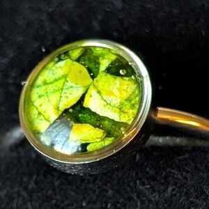 May include: A close-up of a ring with a circular, clear setting. The setting contains a vibrant green and yellow leaf design. The band is gold-colored, and the background is black.