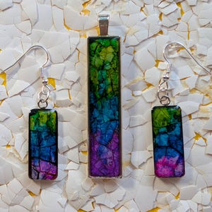May include: A jewelry set featuring a pendant and matching earrings. The rectangular pieces display a vibrant blend of green, blue, and purple hues, encased in silver-toned metal. The earrings have silver hooks. The set is displayed on a background of white fragments.