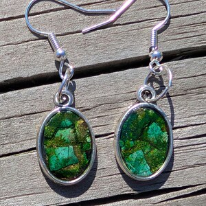 May include: A pair of silver drop earrings. Each earring has an oval, green and gold stone set in a silver frame. The earrings have a hook closure. The earrings are suitable for pierced ears.