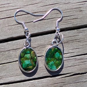 Puede incluir: Pendientes plateados con piedras ovaladas moteadas en verde y dorado. Los pendientes tienen un cierre de gancho y se muestran sobre una superficie de madera. Las piedras tienen un aspecto de mosaico.