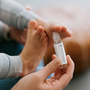 May include: A clear glass bottle with a white label that reads "Calming Baby Blend". The bottle is being held in a person's hand.