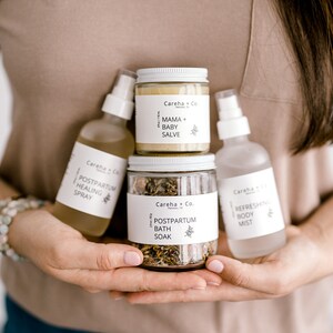 May include: A set of four postpartum care products in white bottles and jars with black text. The products include a healing spray, a bath soak, a body mist, and a mama and baby salve.