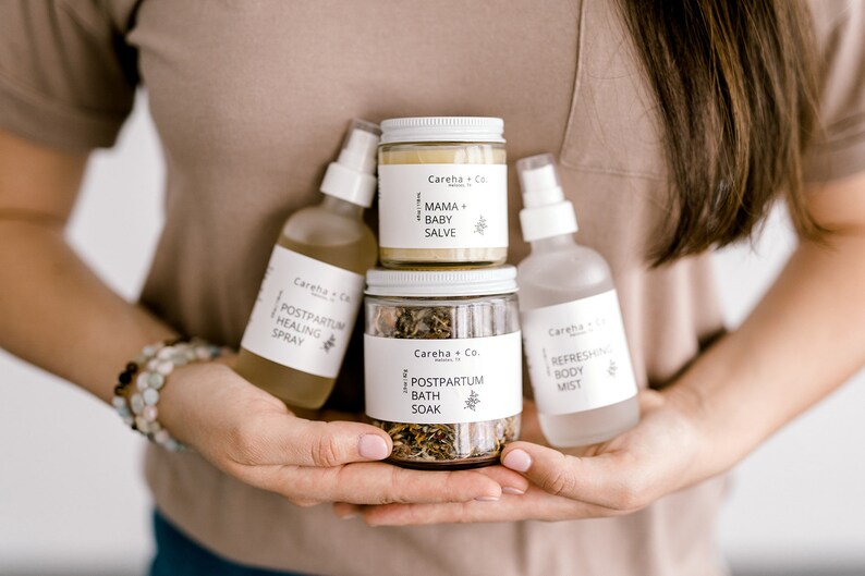 May include: A set of four postpartum care products in white bottles and jars with black text. The products include a healing spray, a bath soak, a body mist, and a mama and baby salve.