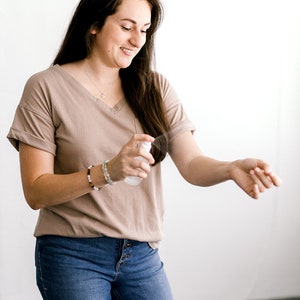 May include: A woman wearing a light brown t-shirt and blue jeans is spraying a clear bottle on her arm.