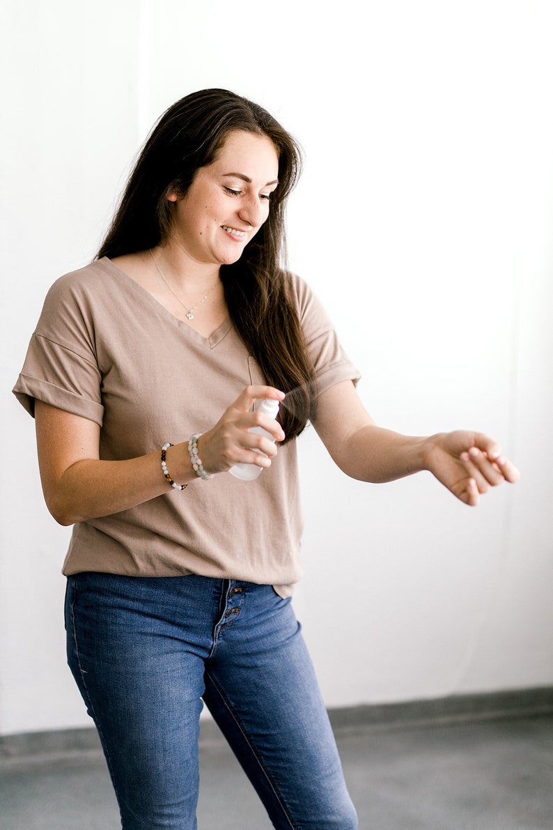 May include: A woman wearing a light brown t-shirt and blue jeans is spraying a clear bottle on her arm.