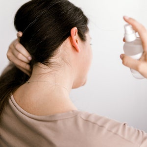 May include: A person with long brown hair is spraying a clear bottle of hair product on their hair.