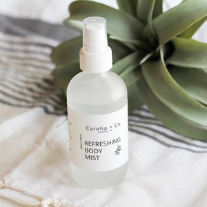 May include: A clear glass bottle with a white spray top and a white label that reads "Careha + Co. Houston, TX Refreshing Body Mist 4 fl oz / 118 ml".
