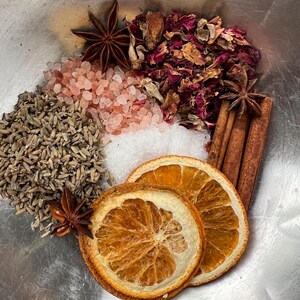 May include: A collection of natural ingredients including dried orange slices, star anise, cinnamon sticks, pink Himalayan salt, lavender buds, rose petals, and white salt. The ingredients are arranged in a metal bowl, showcasing a variety of textures and colors.