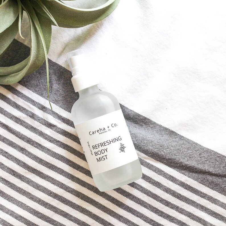 May include: A clear glass bottle with a white spray nozzle and a white label that reads "Careha + Co.  Maridel, TX  Refreshing Body Mist". The bottle is sitting on a white and gray striped fabric.