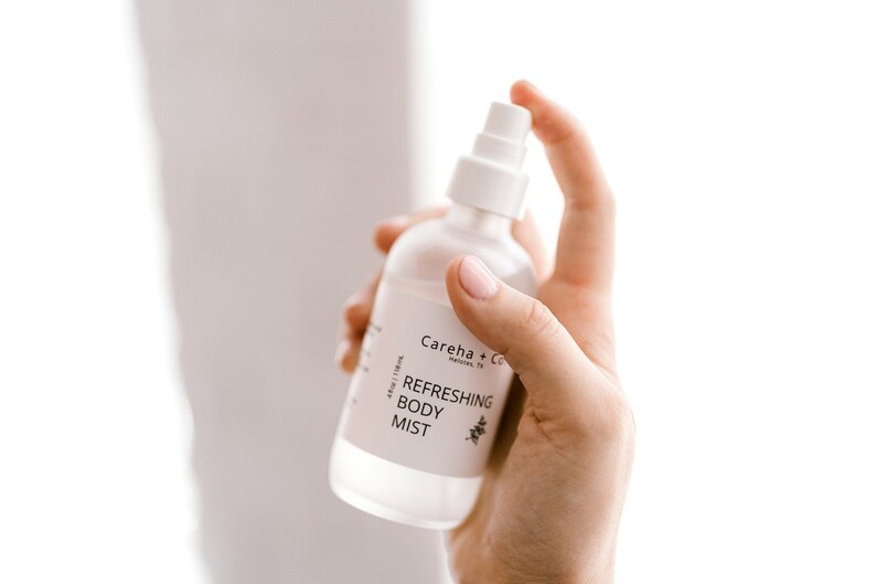 May include: A white bottle of Careha + Co. Refreshing Body Mist. The bottle is clear and has a white label with black text. The bottle is being held by a hand.