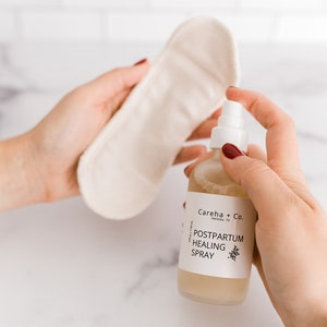 May include: A clear glass bottle with a white label that reads "Careha + Co. Helotes, TX" and "Postpartum Healing Spray". The bottle is being held by a hand with red nail polish.