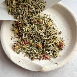 May include: Herbal tea blend being poured onto a white ceramic plate. The tea contains dried green herbs, yellow chamomile flowers, and red rose petals. The plate has a slightly rough texture and a light beige color.