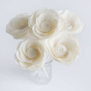 May include: A bouquet of six white wooden flowers with a spiral centre, arranged in a clear glass vase.